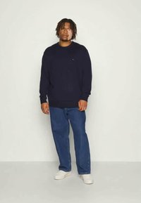 Navy pullover sweater with ribbed cuffs and hem, worn over a black shirt, paired with blue jeans and white sneakers. Simple design, casual fit.