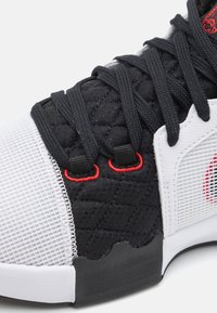 Scarpa sportiva in bianco e nero con texture trapuntata, pannello in rete, dettagli rossi e lacci neri. Presenta una punta arrotondata e suola in gomma.
