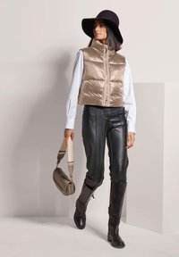 Frau trägt eine beige Daunenweste, ein weißes Hemd, schwarze Lederhosen, schwarze Stiefel und einen breiten Hut, während sie eine beige Handtasche trägt und geht.