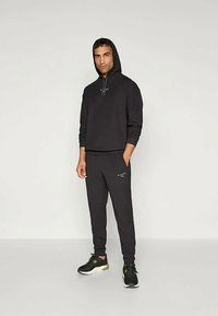 Schwarzer Hoodie mit Kordelzug, kombiniert mit schwarzen Jogginghosen. Verfügt über ein minimalistisches Logo, eine glatte Textur und sportliche Schuhe.