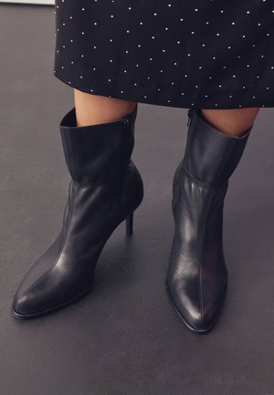Zwarte leren enkellaarzen met puntige neus en stilettohakken, gedragen bij een zwarte rok met kleine witte stippen.