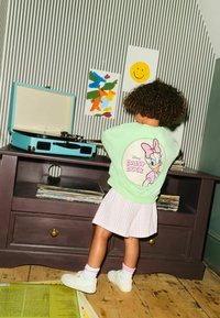 Menta zöld pulóver Daisy Duck grafikával, fehér pliszírozott szoknya, fehér tornacipő, a háttérben csíkos tapéta színes műalkotással.