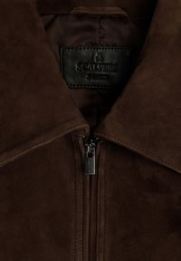 Acercamiento al cuello de una chaqueta de ante marrón con una cremallera centrada y una etiqueta de marca de cuero negro que dice "Scalpers."