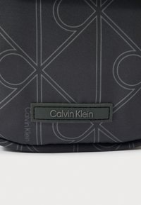 Must kangas peene geomeetrilise mustriga ja ristkülikukujuline must silt, millel on hallid tähed "Calvin Klein".