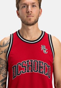 Červený basketbalový dres z jemnej siete s čiernymi akcentmi, odvážnym logom "DCSHOECO", okrúhlym výstrihom a bezrukávovým dizajnom.