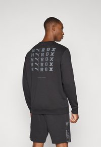 Black sweatshirt with a round neck, featuring a repeated graphic design on the back. Material is soft with a textured finish.