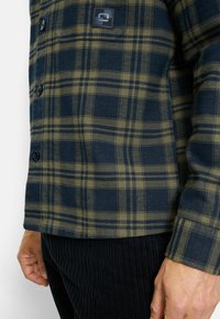 Plaid shirt featuring navy and olive green colors, made of a textured fabric. Includes button closures and a small logo patch on the chest.