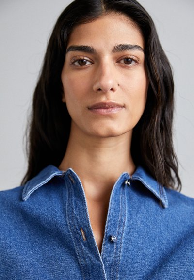 Blå denimskjorte med spiss krage, med kontrasterende sømmer og sølvknapper, som viser en nærbilde av det teksturerte stoffet.