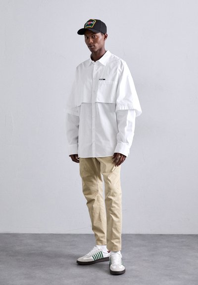 Weißes Hemd mit großen Ärmeln, beiges Hosen und weiße Sneaker mit grünen Streifen. Schwarze Mütze mit buntem Patch-Design.