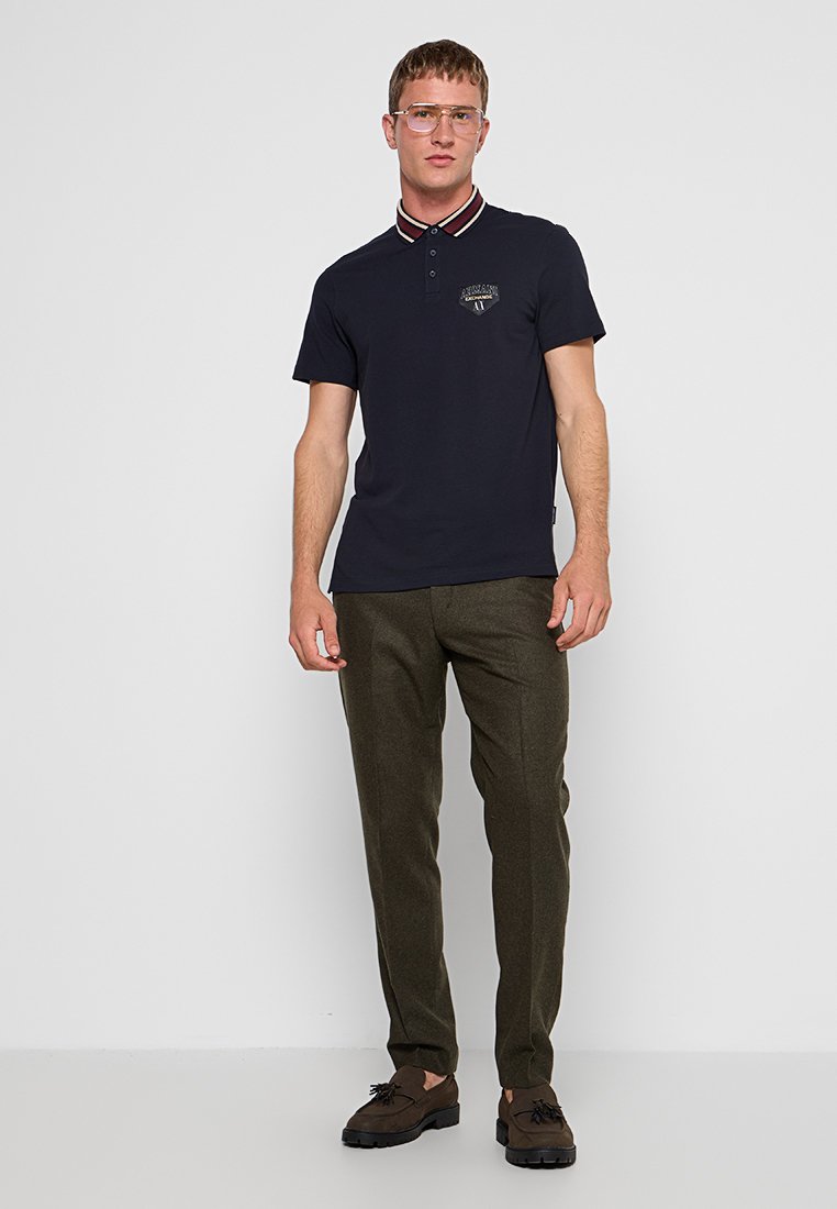 Navy polo shirt met contrasterende rode en witte gestreepte kraag, geborduurd logo op de borst, gecombineerd met olijfgroene broek en bruine schoenen.