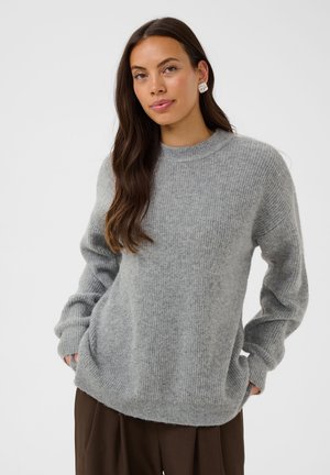 Donna con lunghi capelli castani che indossa orecchini d'argento, un maglione grigio lavorato a maglia e pantaloni marrone scuro, in piedi davanti a uno sfondo chiaro e semplice.
