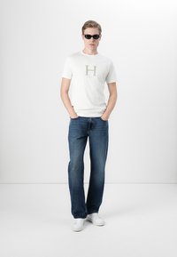 Hvit T-skjorte med grå "H"-logo, blå jeans og hvite joggesko. Modell iført mørke solbriller, står mot en ensfarget bakgrunn.