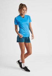 Blå atletisk t-shirt med korte ærmer, parret med blågrønne shorts med et chevron-mønster. Sorte løbesko med hvide accenter.