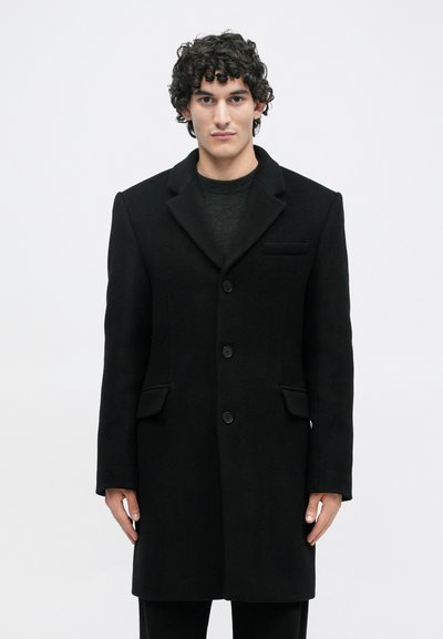 Άνδρας με σγουρά σκούρα μαλλιά ντυμένος με ένα μαύρο παλτό με τρία κουμπιά πάνω από ένα σκούρο πουλόβερ, που στέκεται μπροστά σε έναν απλό λευκό φόντο.