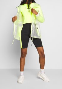 Neon žlutá průhledná bunda do deště s černými šortkami, se stahovací šňůrkou a sportovními botami. Na vršku je logo přes hruď.