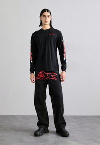 T-shirt nera a maniche lunghe con grafiche rosse sulle maniche. Pantaloni neri con un grande design di coniglio rosso. Scarpe nere spesse. Sfondo semplice.