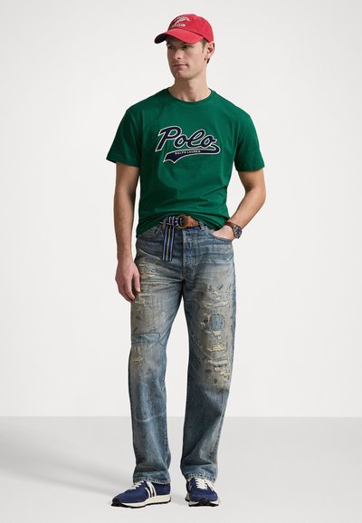 Πράσινο βαμβακερό μπλουζάκι με λογότυπο "Polo", φθαρμένο μπλε τζιν με μπαλώματα και σχισίματα, κόκκινο καπέλο και μπλε αθλητικά παπούτσια με λευκές λεπτομέρειες.