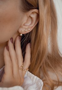 Gouden oorbellen met parels en kristallen accenten, in verschillende vormen. Ringen met gedraaide banden en kleine edelstenen op de vingers, in zacht licht.