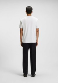 Mężczyzna z krótkimi ciemnymi włosami, ubrany w gładką białą koszulę z krótkim rękawem, czarne spodnie i czarne buty, stojący na jednolitym szarym tle.