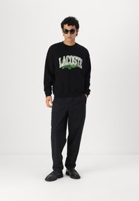 Sudadera negra con logo verde de "LACOSTE" y gráfico de cocodrilo, combinada con pantalones oscuros y zapatos negros. Modelo usando gafas de sol redondas.