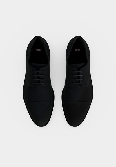 Paire de chaussures habillées en cuir noir avec lacets et détails bout rapporté, vues de dessus sur un fond blanc uni.