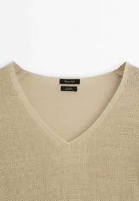 V-kaulus beige neule, jossa on löysä kudontakuvio. Merkit brändistä ja materiaalista (100 % pellava) näkyvät kaulassa.
