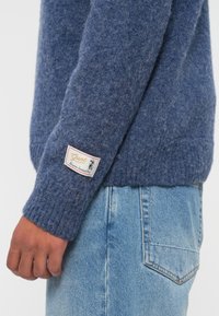 Maglione blu a maglia con polsini a costine, dotato di etichetta del marchio sulla manica. Abbinato a jeans di denim blu chiaro.