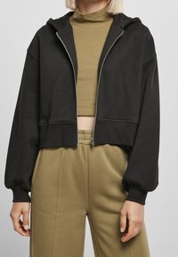 Sudadera negra recortada con cremallera frontal, mangas extragrandes y puños acanalados, combinada con una camiseta verde oliva y pantalones a juego.