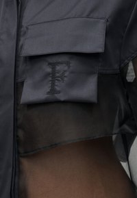 Μαύρο κοντό σακάκι με μεγάλη τσέπη με καπάκι που φέρει ραμμένο το γράμμα "F" πάνω σε διαφανές μαύρο τοπ.