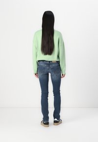 Sweatshirt oversize vert, jeans skinny bleus, vue de dos. Cheveux longs et foncés. Baskets avec des rayures blanches et noires et semelle marron clair.
