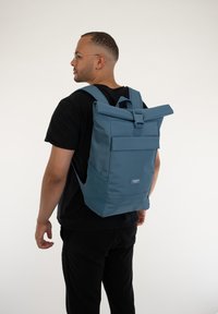 Kék roll-top hátizsák textúrált felülettel, állítható pántokkal, elülső zsebbel és csatos záródással. Simaszerű anyag minimális márkázással.