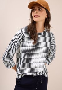 Junge Frau mit braunen Haaren und Sommersprossen trägt eine braune Mütze, einen schwarz-weißen gestreiften Pullover und eine marineblaue Hose. Sie schaut zur Seite mit den Händen hinter dem Rücken.