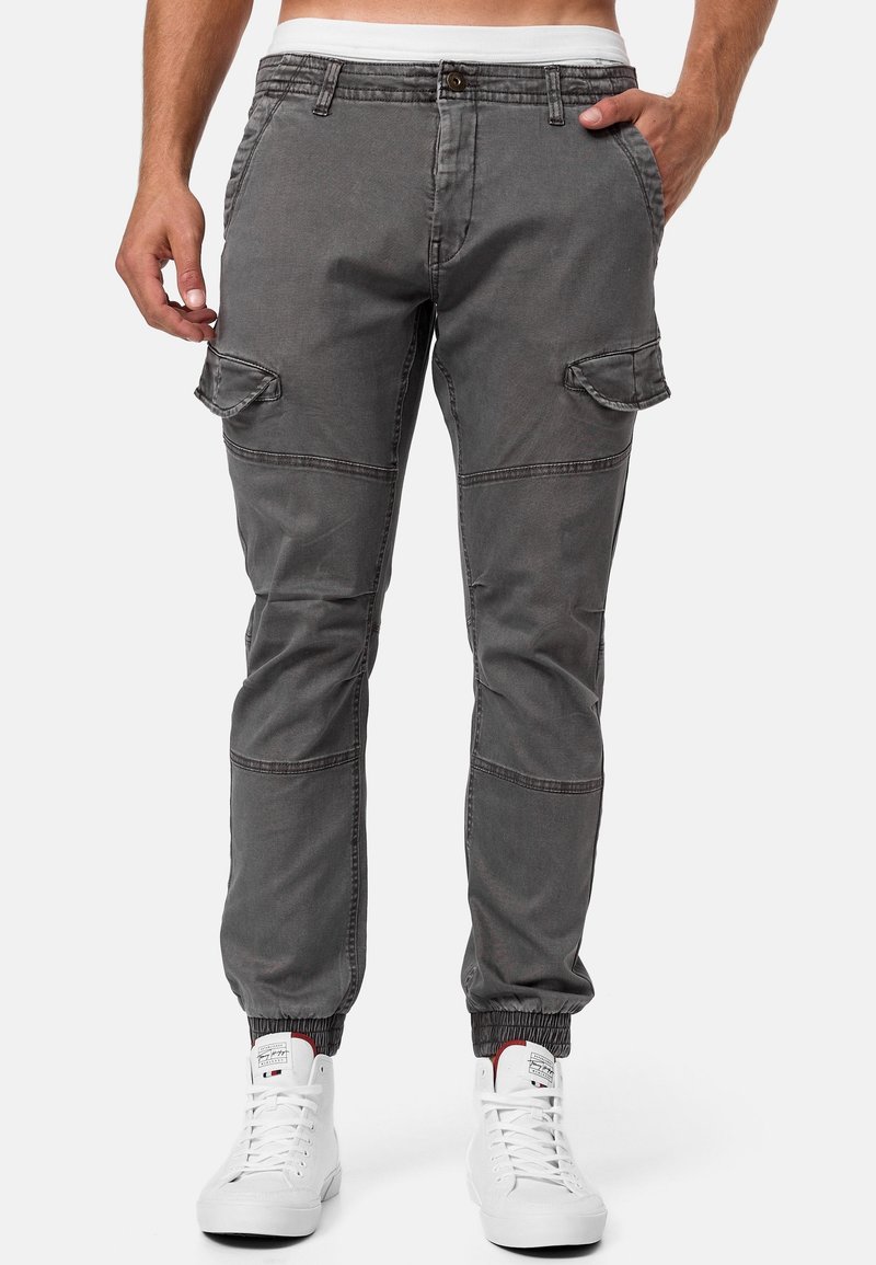 Homme portant un pantalon cargo gris avec poches à rabat et ourlets élastiques, associé à des baskets montantes blanches, les mains dans les poches.