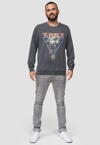 Grijs sweatshirt met lange mouwen met een T. Rex grafisch ontwerp binnen een blauwe driehoek, gecombineerd met lichtgrijze jeans en witte sneakers.