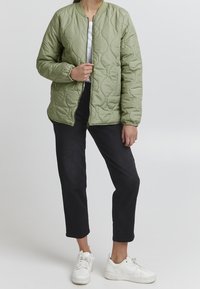 Pikowana zielona kurtka bomberka z zamkiem na przodzie, ściągaczami na rękawach i luźnym krojem, w połączeniu z ciemnymi spodniami i białymi sneakersami.