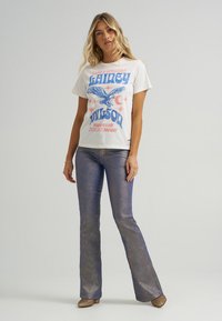 Gaiša krāsa grafiskā T-kreklā ar zilu un rozā drukājumu, apvienota ar mirdzošiem ziliem platiem biksēm un zelta apaviem.