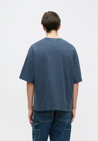 Osoba s krátkými hnědými vlasy, oblečená v modrém oversized tričku a modrých denimových cargo kalhotách, stojící zády k obrazovce na jednobarevném pozadí.