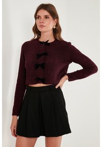 Burgundský texturovaný zkrácený svetr s dlouhými rukávy, který má jako zapínání tři černé sametové mašle, spojený s černou plisovanou mini sukni.