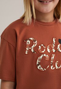 Rūsu krāsas t-krekls ar izšūtām ziedu "Rodeo Club" uzraksta detaļām kontrastējošā krēmu un zaļā krāsā, ar īsām piedurknēm un apaļu kakla izgriezumu.