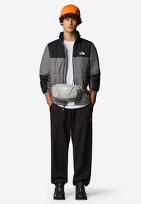 Lichtgewicht, puffy herenjack in zwart en grijs, oranje gebreide muts, heuptas met gaasdetails, zwarte joggers en stevige zwarte sneakers.