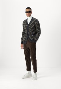 Mørkegrå V-hals cardigan med tan argyle mønster, sorte solbriller, hvid turtleneck, brune fløjl bukser og hvide sneakers.