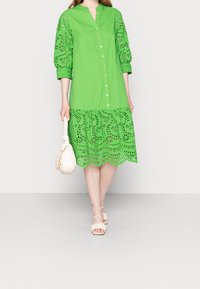 Mujer vestida con un vestido verde abotonado con mangas y dobladillo de encaje, sosteniendo un bolso blanco y usando sandalias beige.