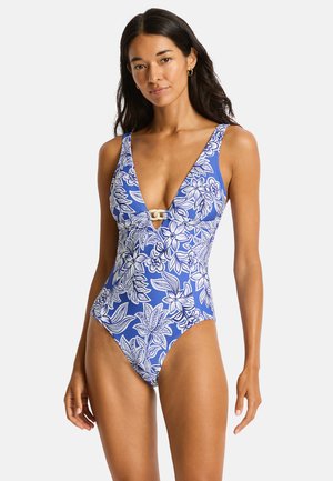 Vrouw die een blauw badpak met witte bloemenprint en diepe V-hals draagt, staand tegen een effen lichte achtergrond.