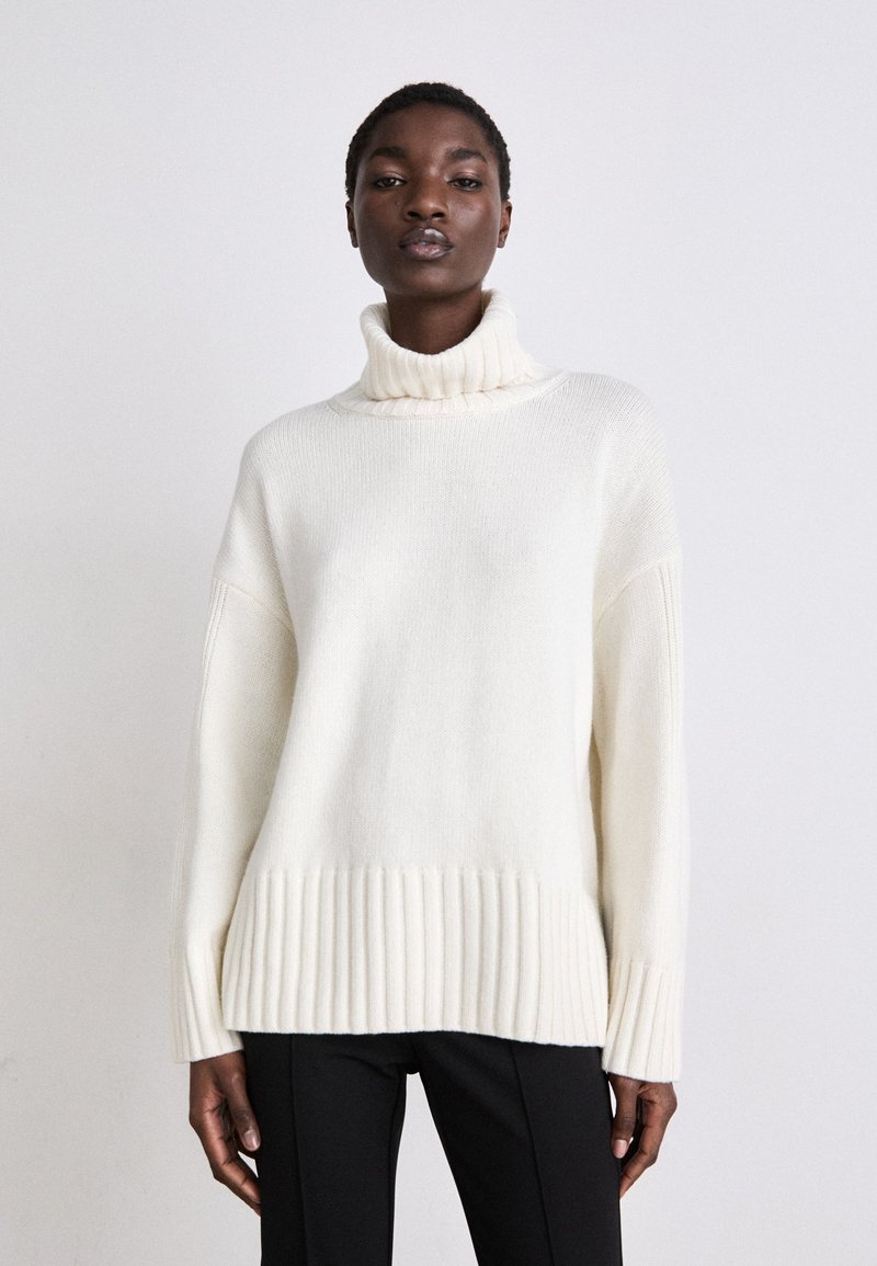 Cremfarbener Rollkragenpullover mit geripptem Material am Saum und den Ärmeln. Verfügt über eine lockere Passform und weiches Strickmaterial. Kombiniert mit schwarzen Hosen.