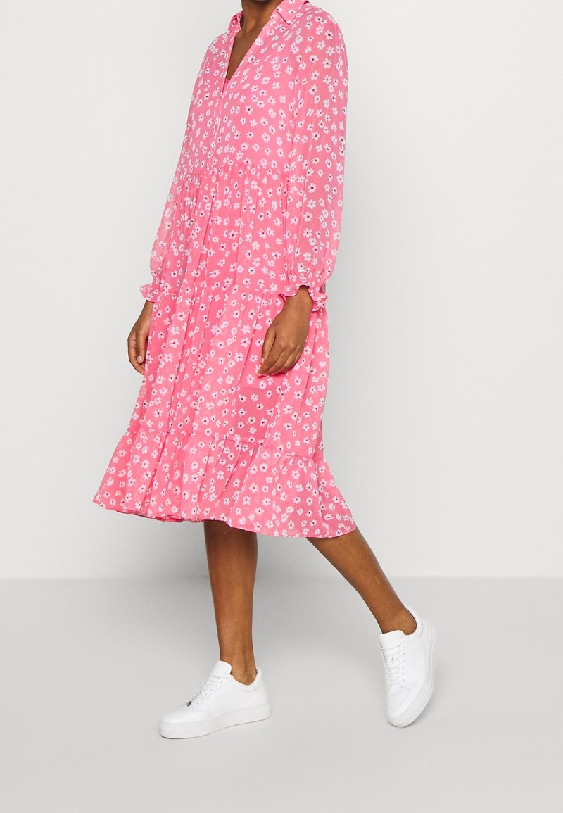 Personne portant une robe midi rose à manches longues avec imprimé floral blanc et des baskets blanches, debout devant un fond clair uni.