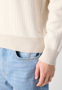 Cremfarbener, gerippter Strickpullover mit strukturierter Verarbeitung, ausgestattet mit einem kontrastierenden, gerippten Saum und Bündchen. Markenlogo-Tag ist an der Seite sichtbar.