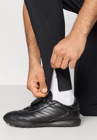 Crne trenirke imaju patentni zatvarač na gležnju. Ruka vuče nogavicu, otkrivajući bijelu čarapu i crnu sportsku obuću.