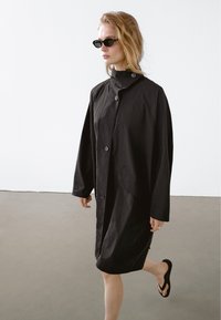 Černý kabát do délky kolen s vysokým límcem, velkými knoflíky a volným střihem. Nosený přes jednoduché černé šaty a žabky. Minimalistický design.