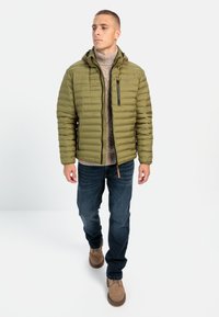 Olivenfarvet pufferjakke med vandret syning, lynlåslomme og justerbar hætte, båret over en beige turtleneck og blå jeans.