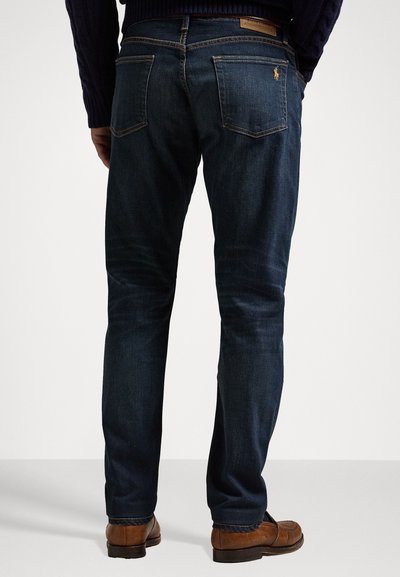 Mørkeblå denimjeans med rett benpassform, med et lærlogo-patch på midjen og to baklommer. Stilt med brune sko.